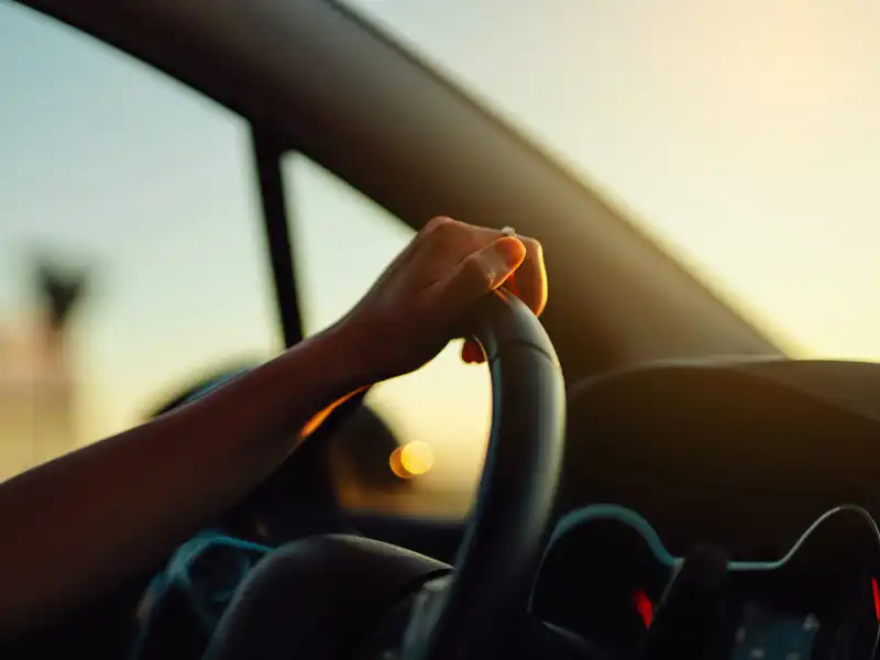 Female hand holding steering wheel in a car during a drive at sunset.