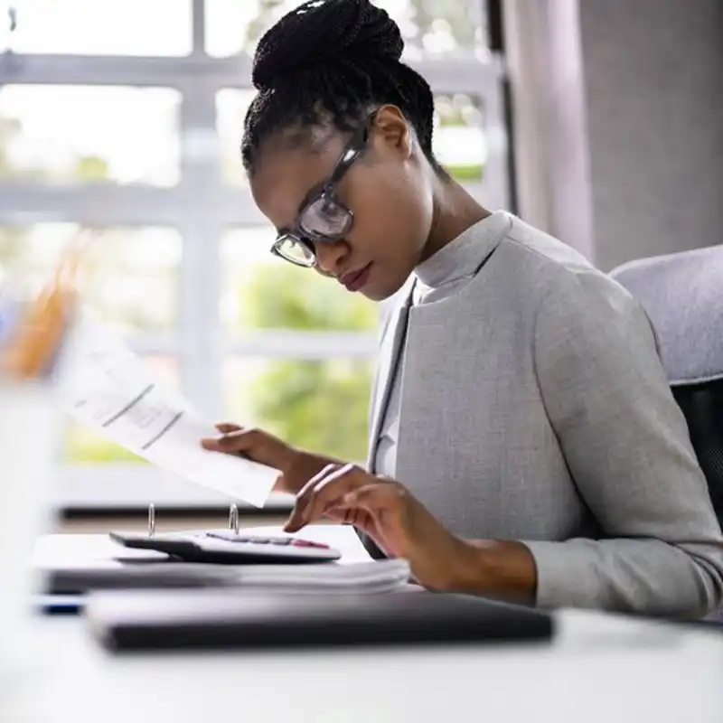 Woman auditor sitting at a desk looking over paperwork with a calculator.