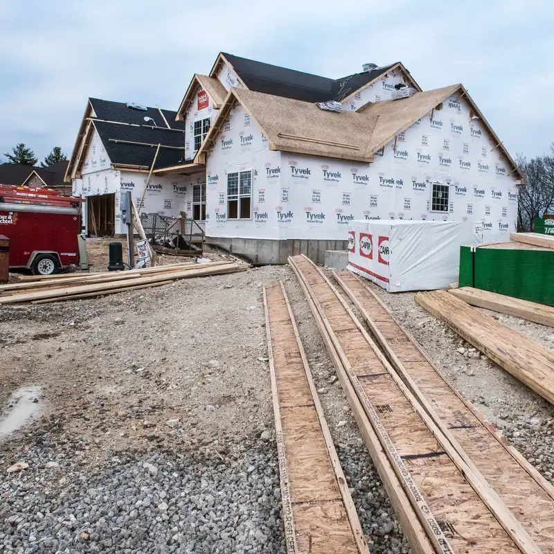 Lumber and I-beams stacked in foreground with new residential home being constructed in background wrapped in Tyvek and work trailer out front,