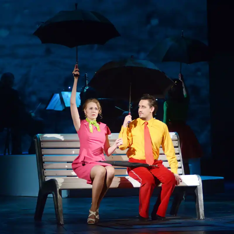 Guy with a girl sit under the umbrellas on a bench during a theatre production.
