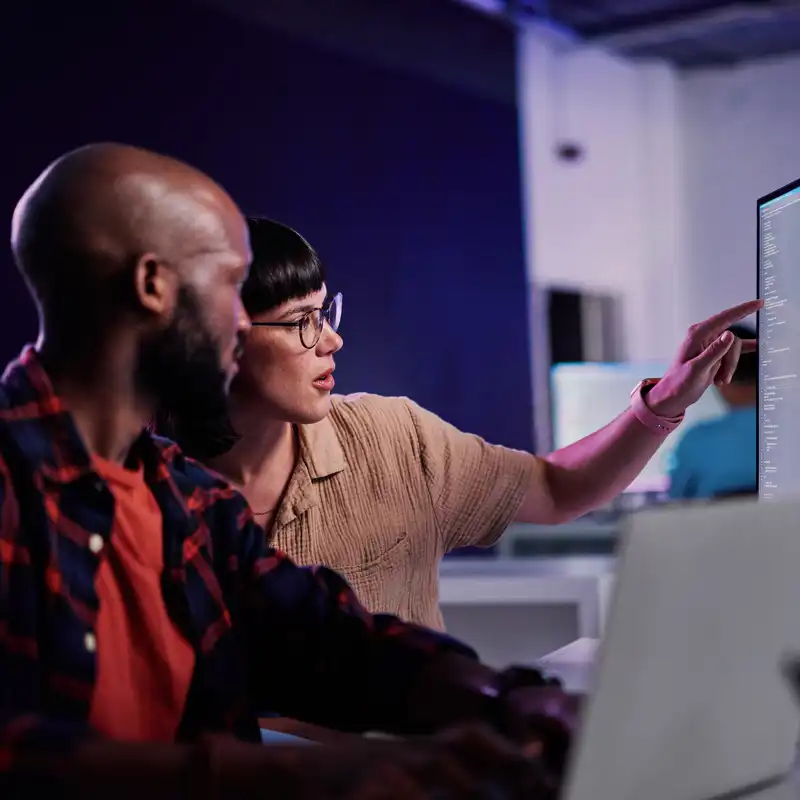 Two people in casual dress study data on a computer monitor.