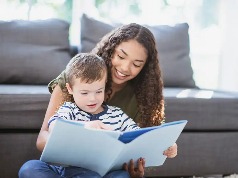 A smiling young woman looks after a child by sharing a book with him.