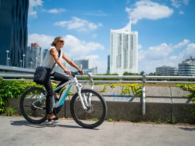A woman with tied hair, sunglasses, and a crossbody cycling on a sunny day with tall buildings in the background.