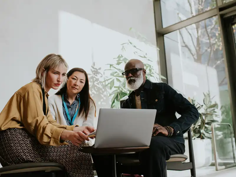 Two women and a man collaborating and discussing while viewing a laptop together.