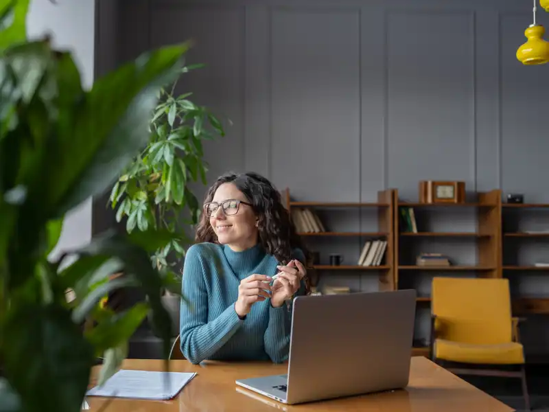 Woman wearing glasses sits at a computer and looks outside her office window.